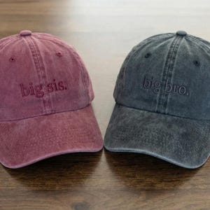 以下が含まれることがあります： 2つの野球帽が表示されています。1つはバーガンディ色、もう1つはチャコールグレーです。バーガンディ色の帽子には「big sis.」と刺繍され、グレーの帽子には「big bro.」と書かれています。どちらの帽子も木製の表面に置かれています。