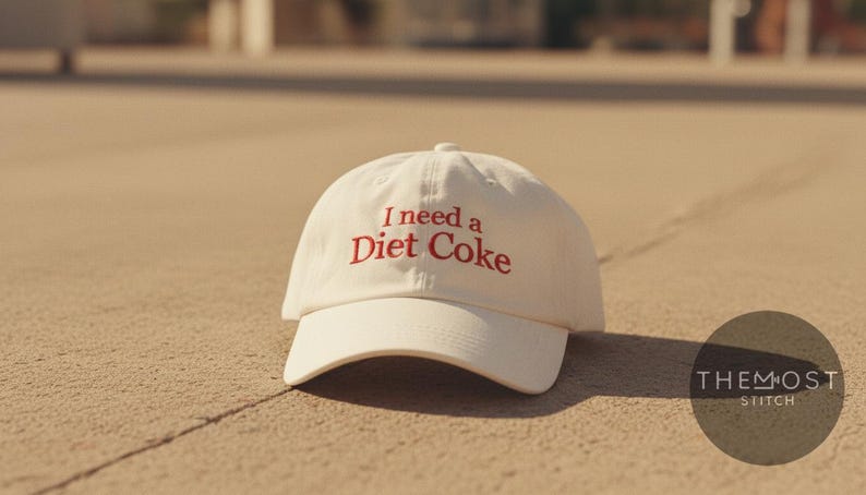 May include: A white baseball cap with a curved brim. The cap features the embroidered text "I need a Diet Coke" in red. The cap is sitting on a light brown surface.