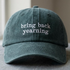 以下が含まれることがあります： 白い文字で「bring back yearning」と刺繍された濃いティール色の野球帽。 つばがカーブしており、布製のストラップ留めが付いています。 帽子は石の表面に置かれています。