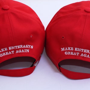 May include: Two red baseball caps with the text "MAKE ENTERASYS GREAT AGAIN" embroidered in white. The caps have a curved brim and an adjustable strap.