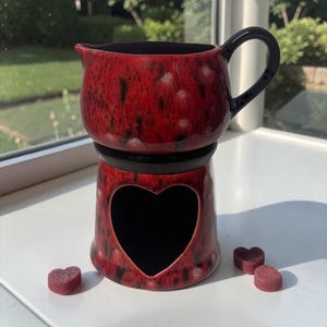 May include: A red and black speckled ceramic wax warmer featuring a pitcher-style top with a handle and a base with a heart-shaped cutout. Three small, dark red heart-shaped wax melts are positioned on the surface near the base of the warmer.
