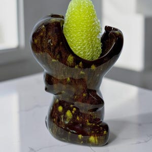 May include: A dark brown ceramic sponge holder with a glossy finish and yellow speckled accents. The holder has an organic, irregular shape with a wide opening at the top, containing a bright lime green, textured, oval-shaped kitchen sponge.