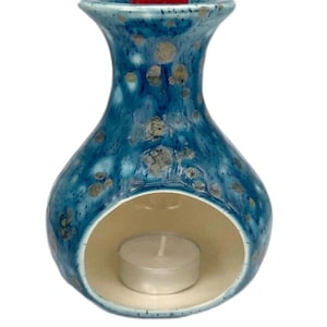 May include: A ceramic oil burner with a glossy blue glaze featuring mottled grey and white spots. The vessel has a wide, rounded base with a large circular opening containing a small white tea light candle and a red stopper at the top.