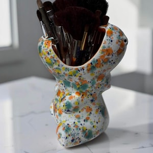 May include: A white ceramic holder shaped like a stylized animal, featuring a speckled pattern of orange, teal, and dark blue paint splatters. The open top contains a collection of various makeup brushes with dark brown and black bristles.