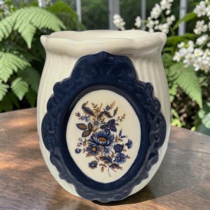 May include: A cream-colored ceramic vessel with a scalloped rim and vertical ribbed texture. The front features a dark blue, ornate, oval-shaped frame surrounding a central cream-colored medallion decorated with a blue and brown floral bouquet.