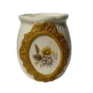 May include: Cream-colored ceramic vase with a gold-colored oval frame. The frame surrounds a floral design with yellow and white flowers. The vase has a textured surface and a ruffled top edge. A decorative home accent.