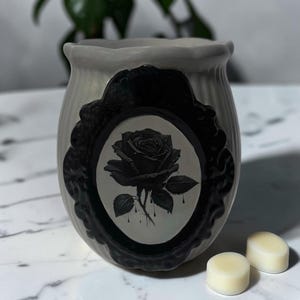 May include: A cream-colored ceramic wax warmer with a fluted exterior and a black oval frame on the front. Inside the frame is a detailed black illustration of a rose with dripping petals. Two small, round, off-white wax melts sit nearby.