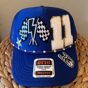 Könnte beinhalten: Blaue Trucker-Mütze mit Mesh-Rückseite und gebogenem Schirm. Die Vorderseite zeigt die weiße Zahl 11 mit goldenem Besatz, karierte Flaggen, einen Blitz und einen Stern. Eine Perlenkette verziert den Schirm. Auf der Mütze steht "OTTO TRUCKER".