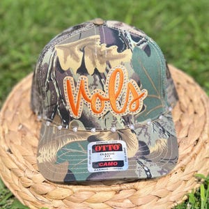 May include: A camouflage baseball cap with the word "Vols" in orange script, embellished with rhinestones. The cap features a mesh back and a curved brim. The cap has a "CAMO" label.