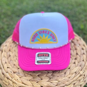 May include: A pink trucker hat with a white front panel featuring a "Good Vibes Only" design in yellow and blue. The hat has a pink brim and mesh back. The hat has a "OTTO" tag.