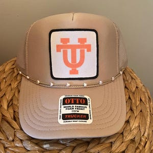 May include: A tan trucker hat with a white and coral logo patch. The hat has a mesh back, a curved brim, and a pearl-like beaded accent. The hat features a label that reads "OTTO TRUCKER".