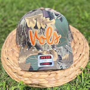 May include: Camouflage baseball cap with the word "Vols" in orange script. The cap has a curved brim and is decorated with small, clear rhinestones. The tag reads "Classic Fit Camo".