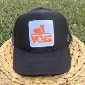 May include: Black trucker hat with a white patch featuring an orange graphic of a person holding a rifle with the text "VOLS" in orange letters.