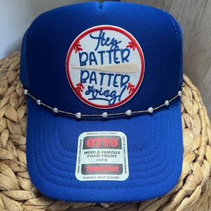 Może przedstawiać: Niebieska czapka typu trucker z naszywką o tematyce baseballowej z napisem "Hey Batter Batter Swing!". Czapka ma perłową opaskę z koralików i zapięcie na zatrzask. Czapka jest wykonana z pianki.