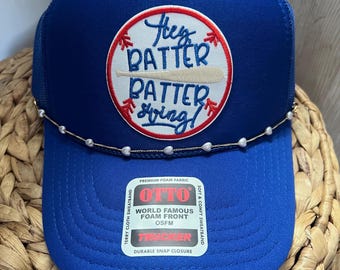 May include: A blue trucker hat with a white and red baseball-themed patch that reads "Hey Batter Batter Swing!" The hat has a pearl-like beaded band and a snap closure. The hat is made of foam fabric.