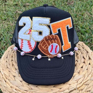 May include: Black trucker hat with a white and gold "25" and an orange "T" on the front. A baseball glove and baseball are also on the front of the hat. A gold chain with white baseball charms hangs from the brim of the hat.