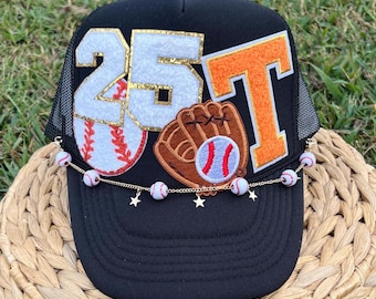 May include: Black trucker hat with a white and gold "25" and an orange "T" on the front. A baseball glove and baseball are also on the front of the hat. A gold chain with white baseball charms hangs from the brim of the hat.