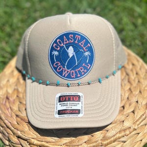 May include: A tan trucker hat with a blue and pink "Coastal Cowgirl" patch. The hat has a turquoise beaded brim and a "World Famous Foam Front" label.