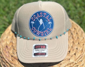 May include: Tan trucker hat with a blue and pink "Coastal Cowgirl" patch. The hat features a turquoise beaded brim and a "World Famous Foam Front" label.