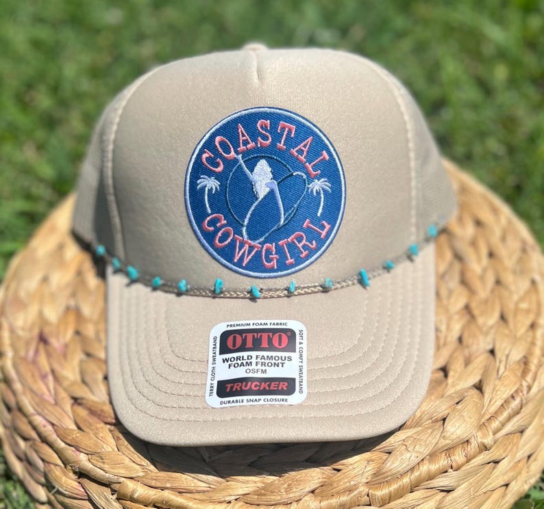May include: Tan trucker hat with a blue and pink "Coastal Cowgirl" patch. The hat features a turquoise beaded brim and a "World Famous Foam Front" label.