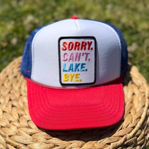 May include: A trucker hat with a white front panel, a red brim, and a blue mesh back. The hat features the text "SORRY. CAN'T. LAKE. BYE." in a colorful, embroidered patch.