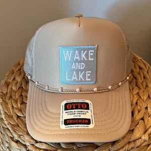 Puede incluir: Gorra de camionero beige con parte trasera de malla y visera curva. La parte delantera presenta un parche cuadrado azul claro con las palabras "WAKE AND LAKE" en blanco. Una hilera de pequeñas cuentas blancas adorna la visera. Cierre a presión.