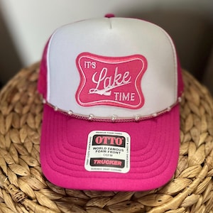 May include: A pink and white trucker hat with the text "It's Lake Time" in a pink, decorative emblem. The hat has a white foam front, pink mesh back, and a curved brim. The hat is OSFM (One Size Fits Most).