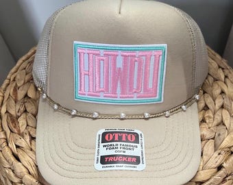 May include: A beige trucker hat with a pearl trim and a pink and turquoise embroidered patch that says "HOWDY". The hat has a black mesh back and a black snap closure. The hat also has a label that says "OTTO WORLD FAMOUS FOAM FRONT OSFM TRUCKER DURABLE SNAP CLOSURE".