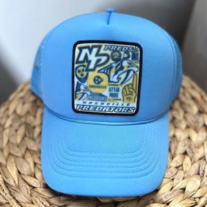 May include: A light blue trucker hat with a black and yellow patch featuring the Nashville Predators logo and the text "Preds" and "Nashville Predators".
