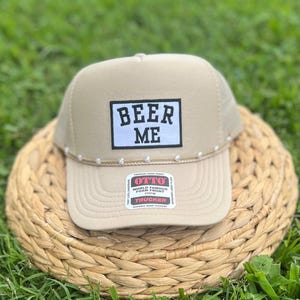 May include: Tan trucker hat with the text "BEER ME" in a black and white box. The hat has a curved brim and a white rope detail along the front. The hat is sitting on a woven basket.