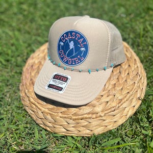 May include: Tan trucker hat with a mesh back and a curved brim. The front features a circular patch with the words "Coastal Cowgirl" and a silhouette of a person surfing. Turquoise beads adorn the brim.