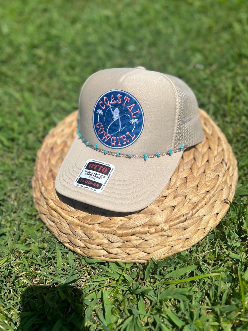 May include: Tan trucker hat with a mesh back and a curved brim. The front features a circular patch with the words "Coastal Cowgirl" and a silhouette of a person surfing. Turquoise beads adorn the brim.