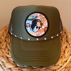 May include: Olive green trucker hat with a black mesh back and a decorative chain. The front features a circular patch with a dog illustration, a stadium, and the letter "T". The hat has a curved brim.