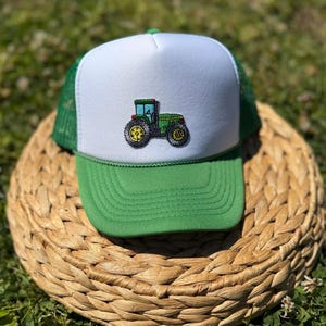 Może przedstawiać: Zielono-biała czapka typu trucker z haftowanym zielonym traktorem i żółtymi kołami. Czapka ma zielony daszek i siatkowy tył oraz biały panel przedni. Wzór traktora jest wyśrodkowany na przednim panelu.