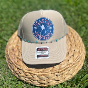 May include: Tan trucker hat with a blue and pink "Coastal Cowgirl" patch. The hat has a curved brim and a row of turquoise beads along the bottom edge. The hat is sitting on a woven basket.