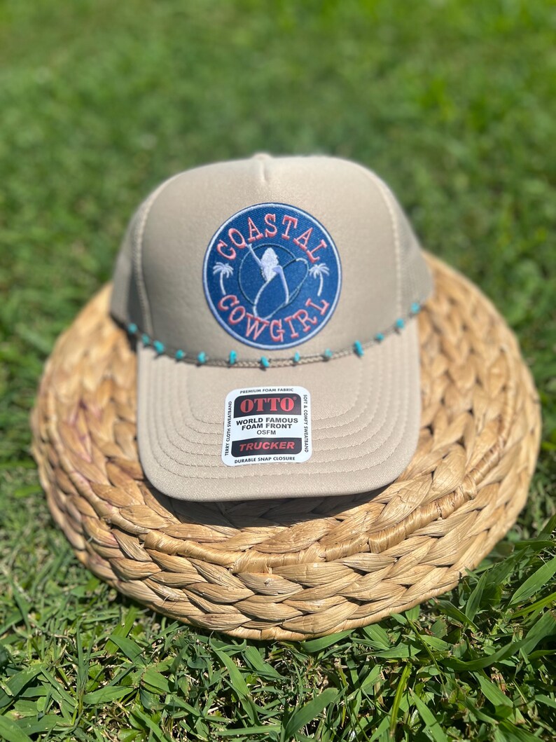 May include: Tan trucker hat with a blue and pink "Coastal Cowgirl" patch. The hat has a curved brim and a row of turquoise beads along the bottom edge. The hat is sitting on a woven basket.