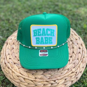 May include: A green trucker hat with a white patch that reads "BEACH BABE" in turquoise lettering. The hat has a curved brim and a beaded accent along the front. The hat is sitting on a woven surface.