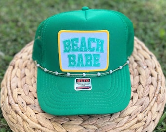 Op de afbeelding: Groene truckerpet met een wit label met de tekst "BEACH BABE" in turquoise. De pet heeft een gebogen klep en een kralenrand. De pet ligt op een geweven oppervlak.