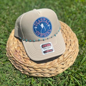 May include: Tan trucker hat with a blue and coral "Coastal Cowgirl" patch. The hat features a mesh back, a curved brim, and a turquoise beaded accent along the brim. The hat is sitting on a woven basket.