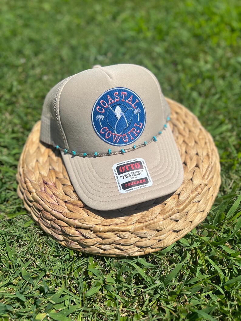 May include: Tan trucker hat with a blue and coral "Coastal Cowgirl" patch. The hat features a mesh back, a curved brim, and a turquoise beaded accent along the brim. The hat is sitting on a woven basket.