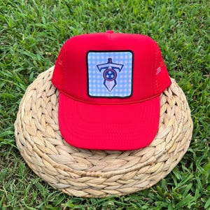 May include: Red trucker hat with a blue and white gingham patch featuring a sword and stars. The patch is a Tennessee Titans logo.