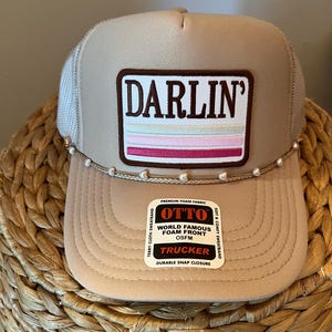 May include: A tan trucker hat with a mesh back and a curved brim. The front features a rectangular patch with the word "DARLIN'" and horizontal stripes. A string of pearls is attached to the brim. The hat has a "OTTO" tag.