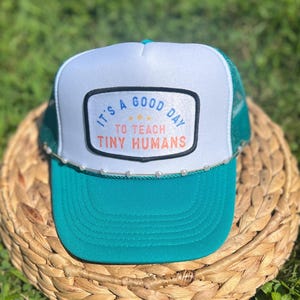 May include: A teal and white trucker hat with a pearl-like trim along the brim. The hat features a rectangular patch with the text "It's a good day to teach tiny humans."