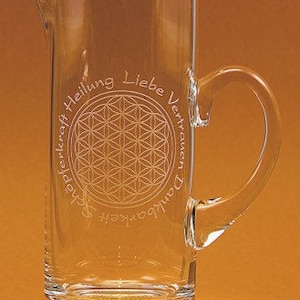 Könnte beinhalten: Ein klarer Glaskrug mit Henkel und Ausguss. Der Krug hat ein geätztes Design eines blütenartigen Musters in einem Kreis, umgeben von Text. Das Wort "handmade" ist am Boden eingeätzt.