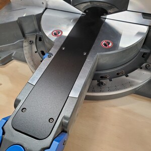 May include: Close-up of a black and silver mitre saw with a blue lever and a red warning sign. The saw has a protractor with markings from 35 to 60 degrees.