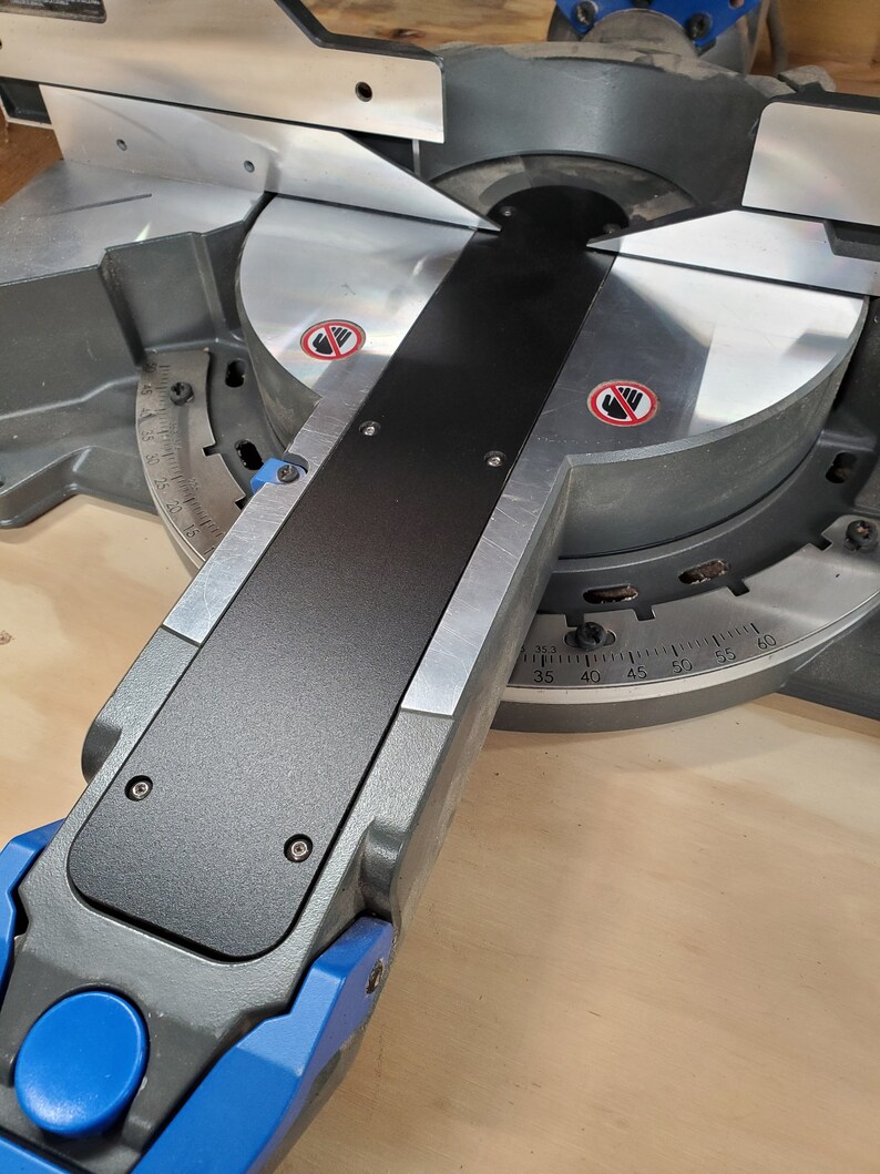 May include: Close-up of a black and silver mitre saw with a blue lever and a red warning sign. The saw has a protractor with markings from 35 to 60 degrees.