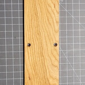 May include: A long, narrow, rectangular wooden plaque with a rounded top and a natural wood finish. The plaque has six evenly spaced holes drilled into it.