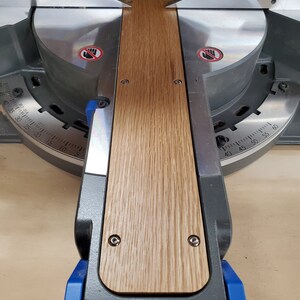 Puede incluir: Primer plano de una sierra de inglete con una pieza de madera que se est&aacute; cortando. La madera es de color marr&oacute;n claro con un patr&oacute;n de veta. La sierra es de color negro y gris con un mango azul. La sierra tiene un transportador con marcas en grados.