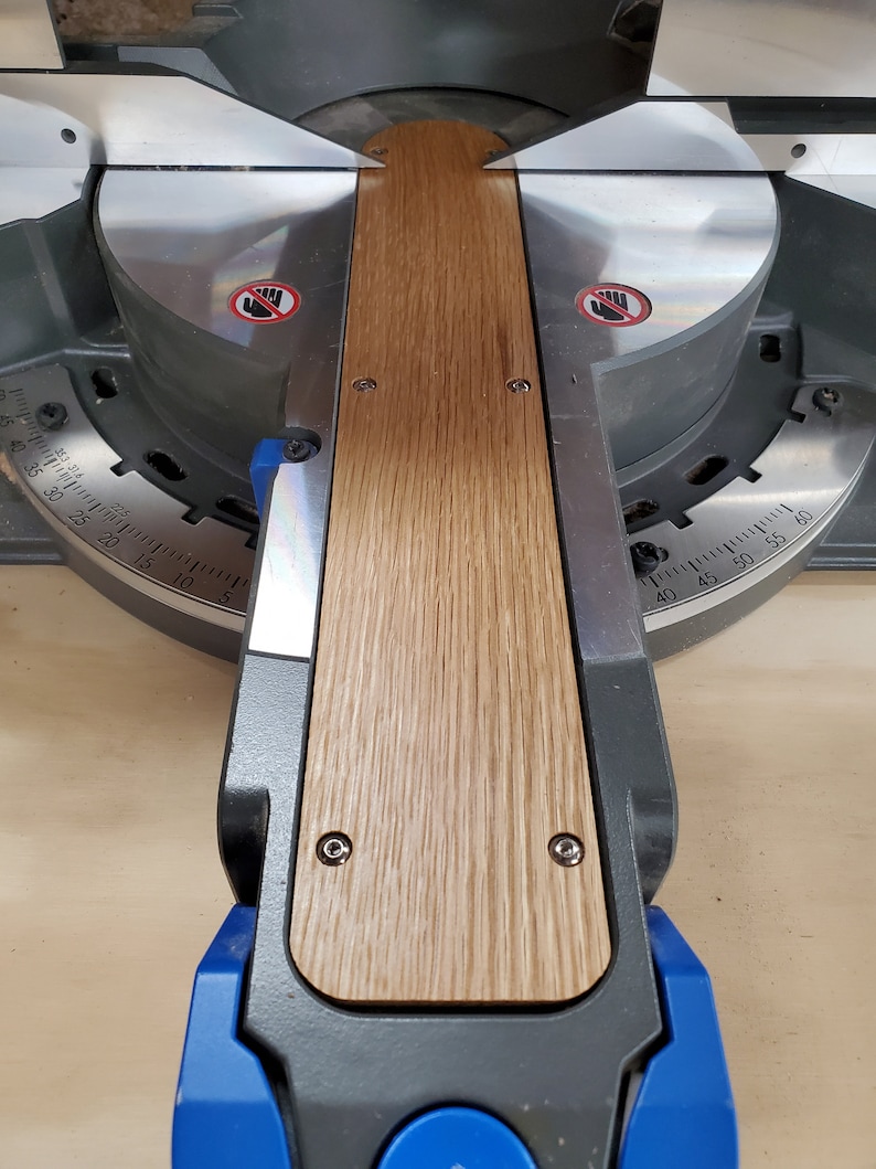 Puede incluir: Primer plano de una sierra de inglete con una pieza de madera que se est&aacute; cortando. La madera es de color marr&oacute;n claro con un patr&oacute;n de veta. La sierra es de color negro y gris con un mango azul. La sierra tiene un transportador con marcas en grados.