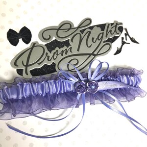 May include: A lavender prom garter with a matching bow and the words "Prom Night" in silver script. The garter features a ruffled tulle design and is adorned with small purple flowers and ribbons. Black high heels are also visible.